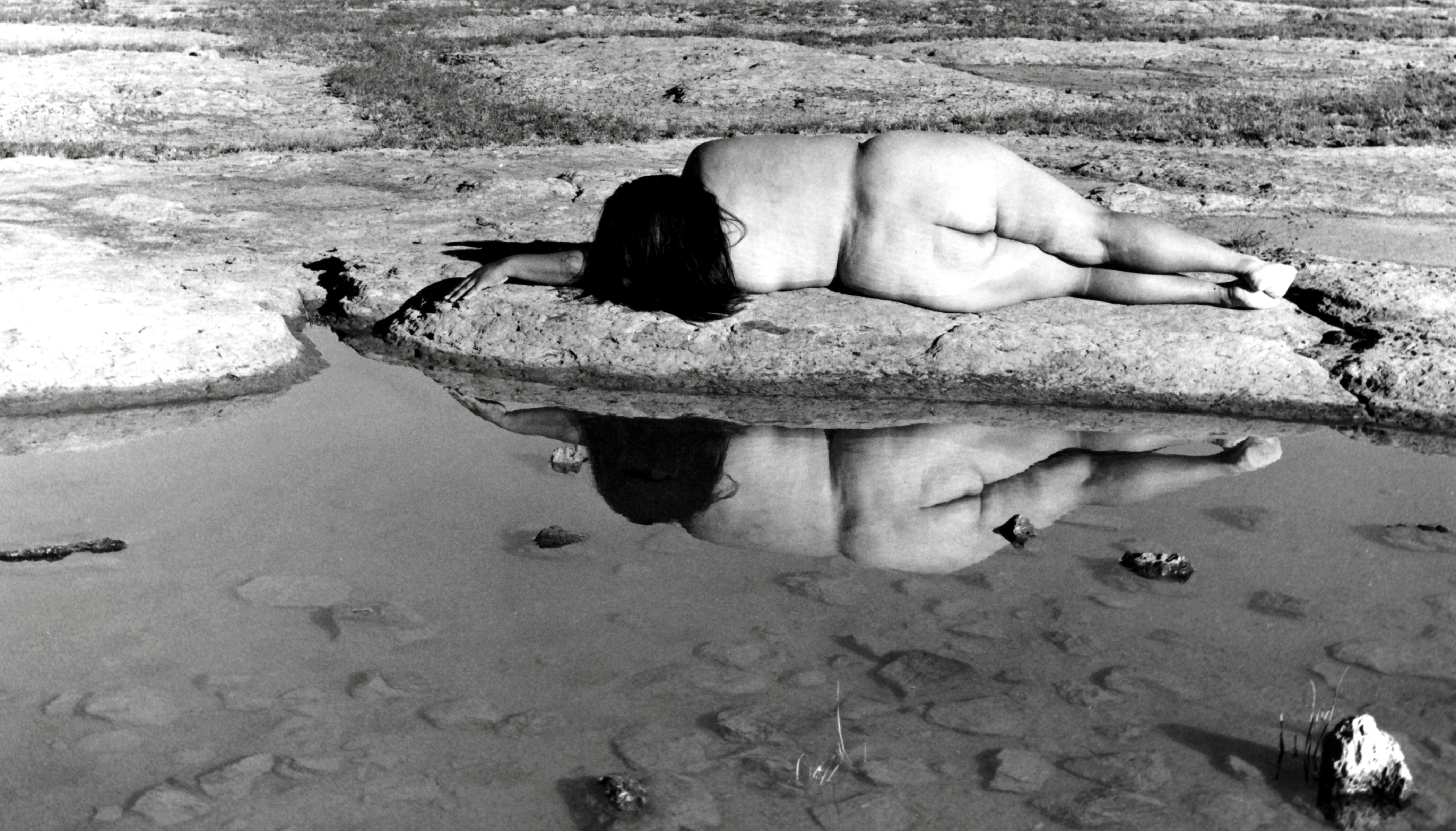 Black and white photograph of Laura Aguilar lying naked on the beach, facing away from viewer towards ocean waves