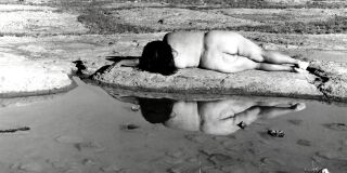 Black and white photograph of Laura Aguilar lying naked on the beach, facing away from viewer towards ocean waves
