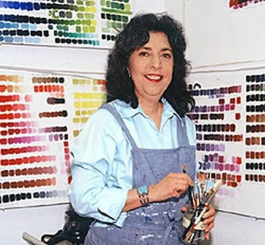A woman in a blue overall holds paint brushes and poses in front of color palettes