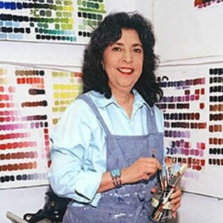 A woman in a blue overall holds paint brushes and poses in front of color palettes