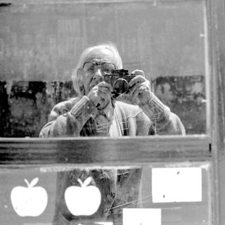 A man with glasses takes a photo of himself in a glass reflection