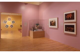 Ocumicho Ceramics in a glass case with framed photographs to the right; artwork displayed in a pink gallery room.