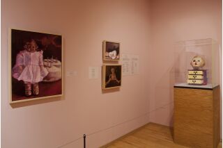 Photo of two gallery walls, with three paintings visible on left wall and glass case against right wall