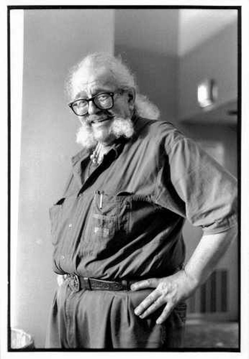 A bearded man with black glasses places his arms at his hips and poses for a photo