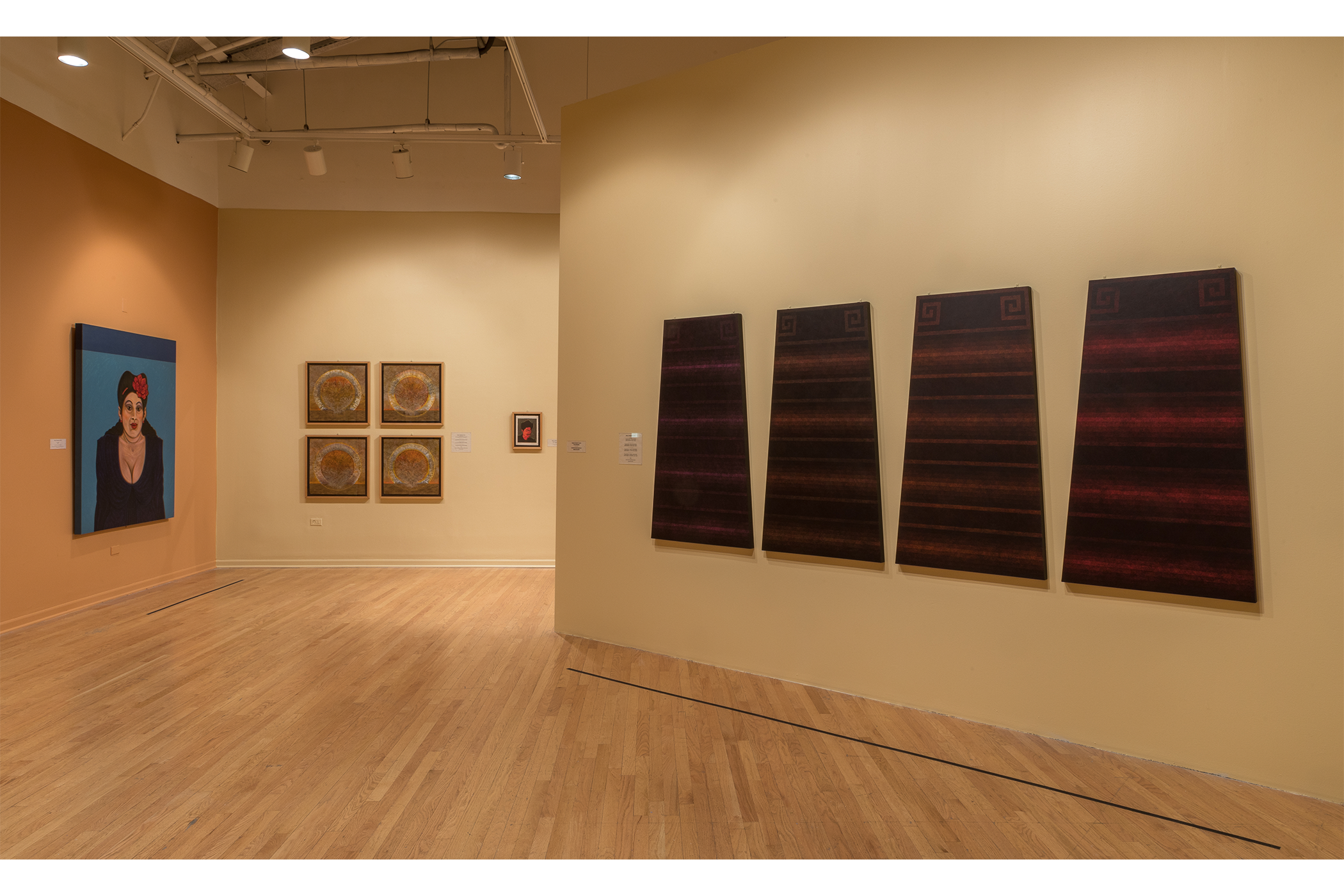 Four artworks hang in the gallery. One, a lady in a black dress, red flower in hair, blue background.  Two, A collection of 4 acrylic painted panels: all with circular emblems in center.  Three, a man with a hat and moustache.  Four, four rectangular shapes, each a different color, and each with horizontal lines running across in varying placements.