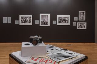 Gallery photo of one wall with five black and white photographs. In the center is a podium with a camera and documents, including a license plate