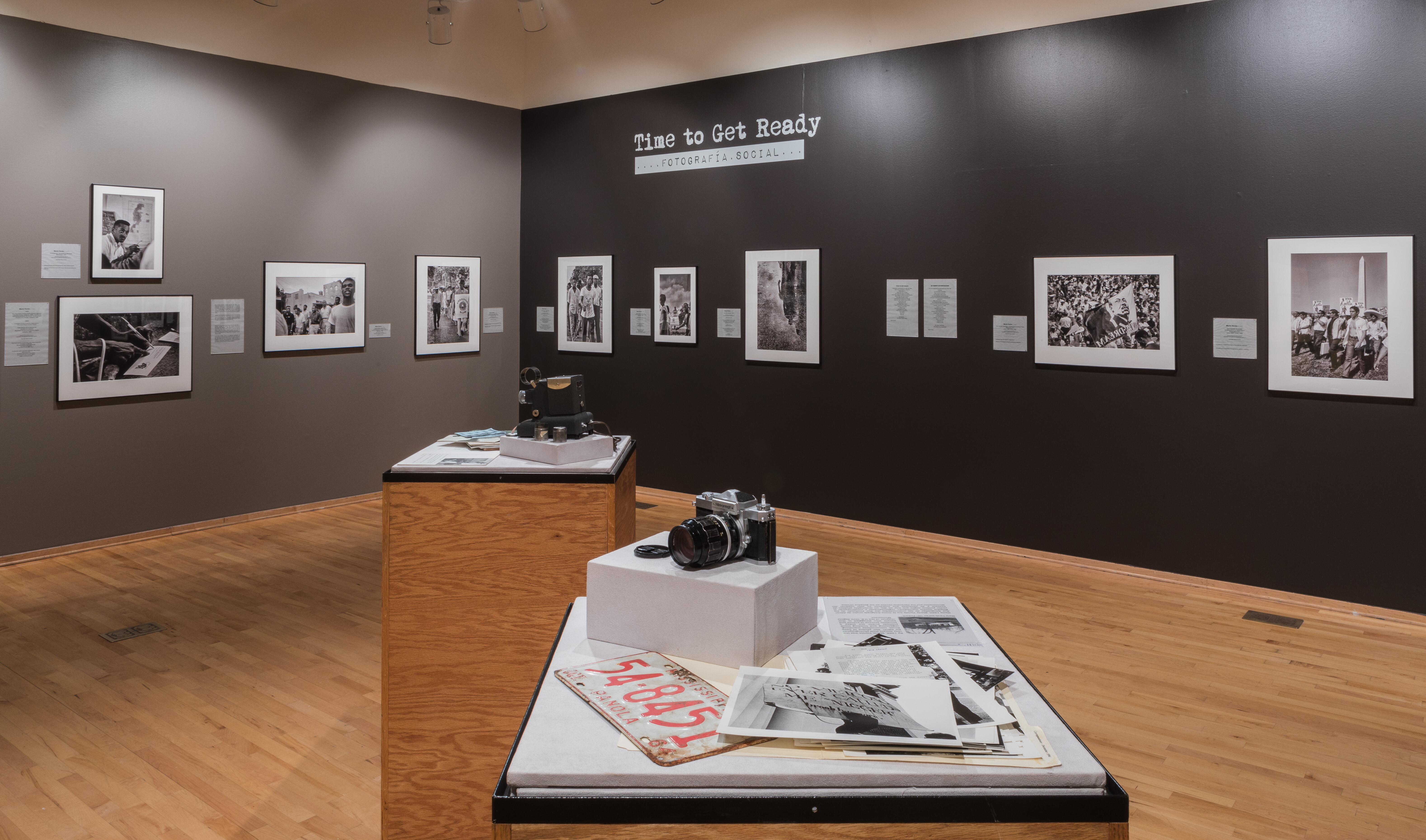 Photo of two gallery walls filled with black and white photos. Two podiums in the center of the room with cameras and paper documents