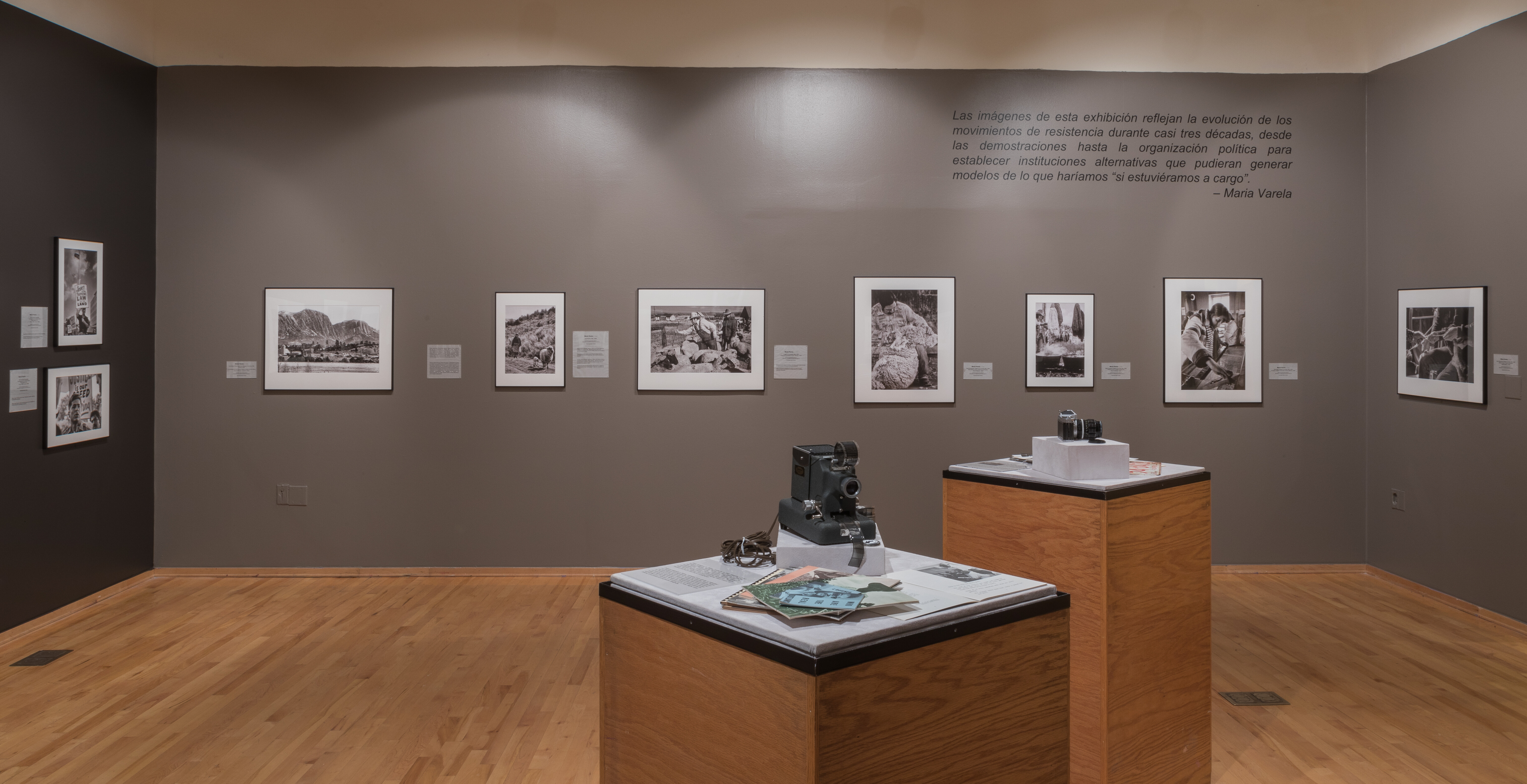 Photo of one gallery wall with six black and white photographs. In the center of the room are two podiums with cameras and paper documents