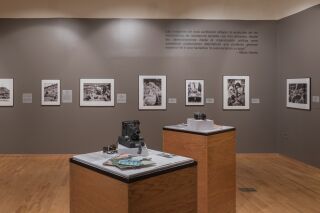 Photo of one gallery wall with six black and white photographs. In the center of the room are two podiums with cameras and paper documents