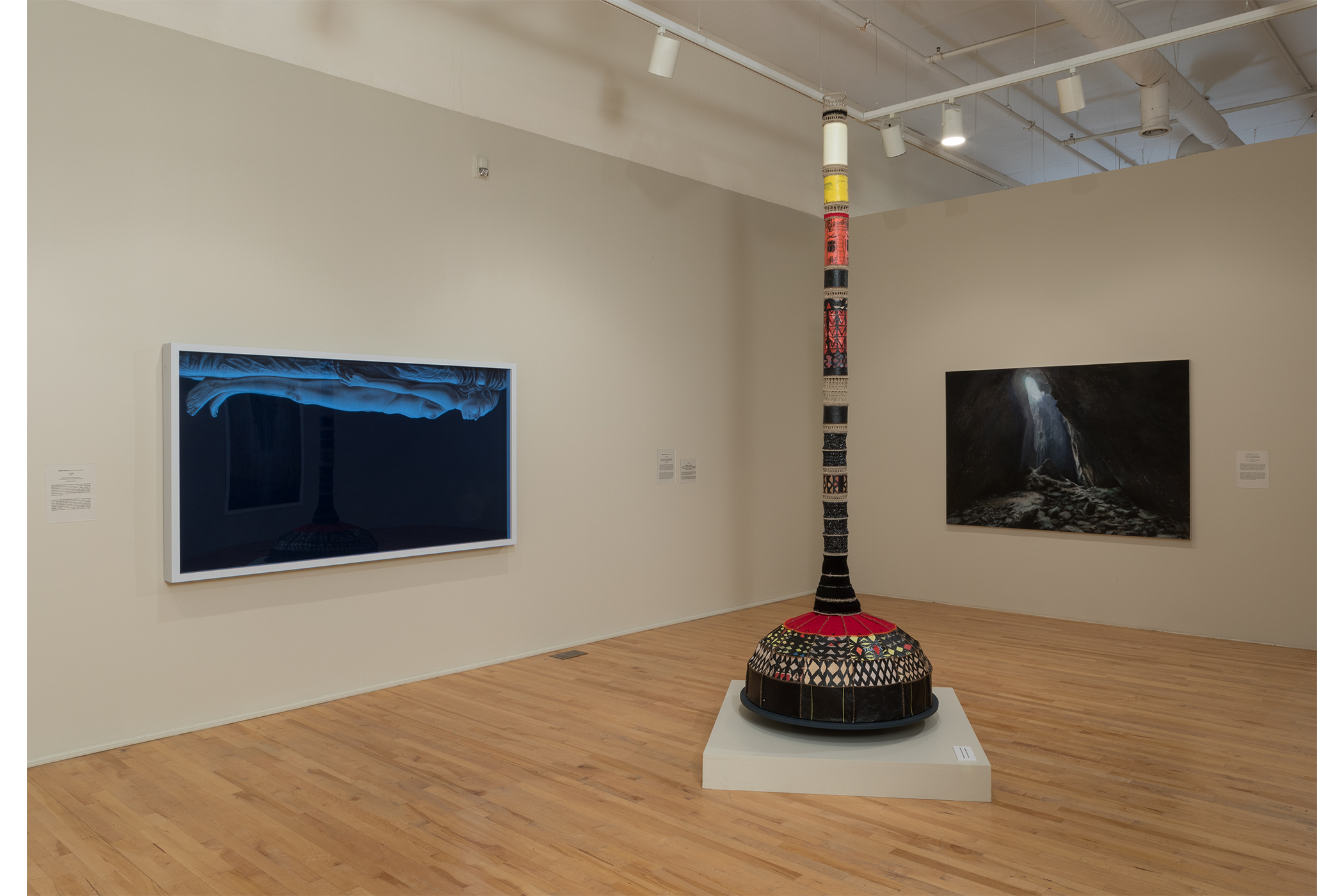 Three artworks on display: man laying flat from the top facing downward, a mixed media pillar, oil painting of a cave opening with light shining through.