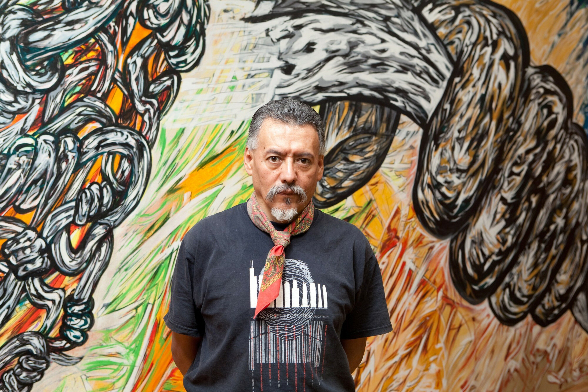 A man in a black shirt and bandana poses before a colorful mural