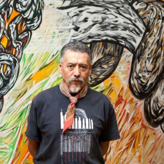 A man in a black shirt and bandana poses before a colorful mural