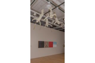 a moving installation hangs from the ceiling, of four people with their arms spread wide, each with a few strands of ribbon from their heads