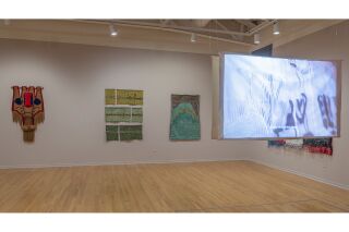 Three woven pieces visible on wall with hanging video screen hanging from ceiling on right side