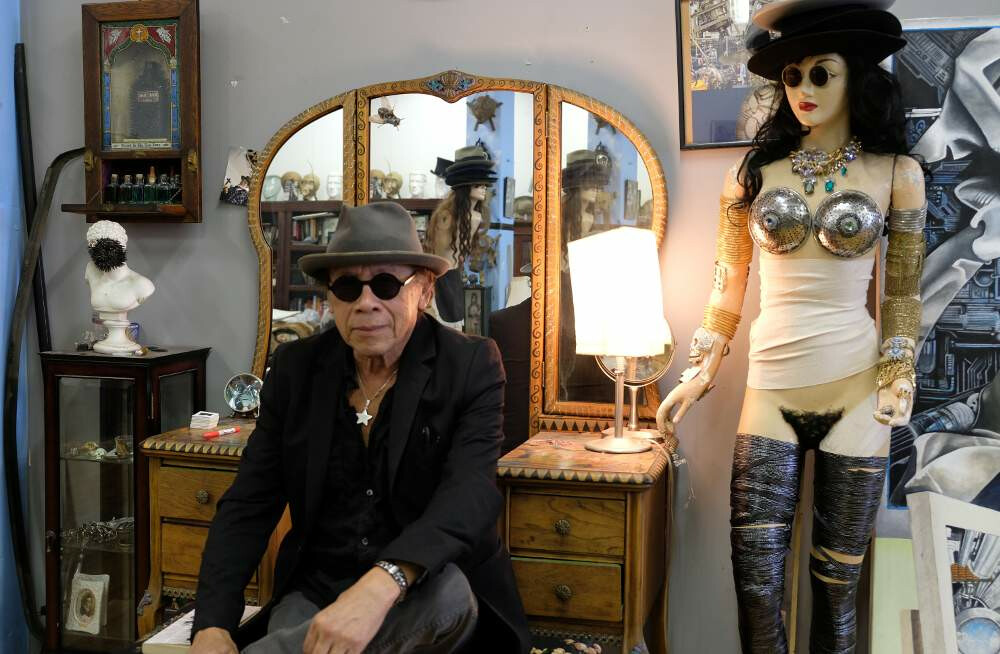 A man in a hat, dark glasses, and a black jacket sits in front of a vanity and next to a mannequin for a photo