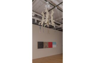 The installation hangs from the ceiling, currently stationary, four people with their arms spread wide, each with a few strands of ribbon from their heads