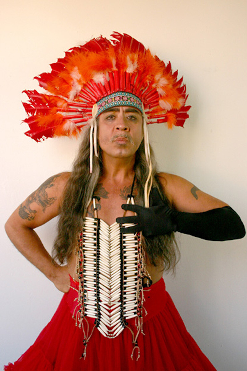 A peron wearing a headpiece holds one arm at their hip and another arm in a long black glove in front of their chest