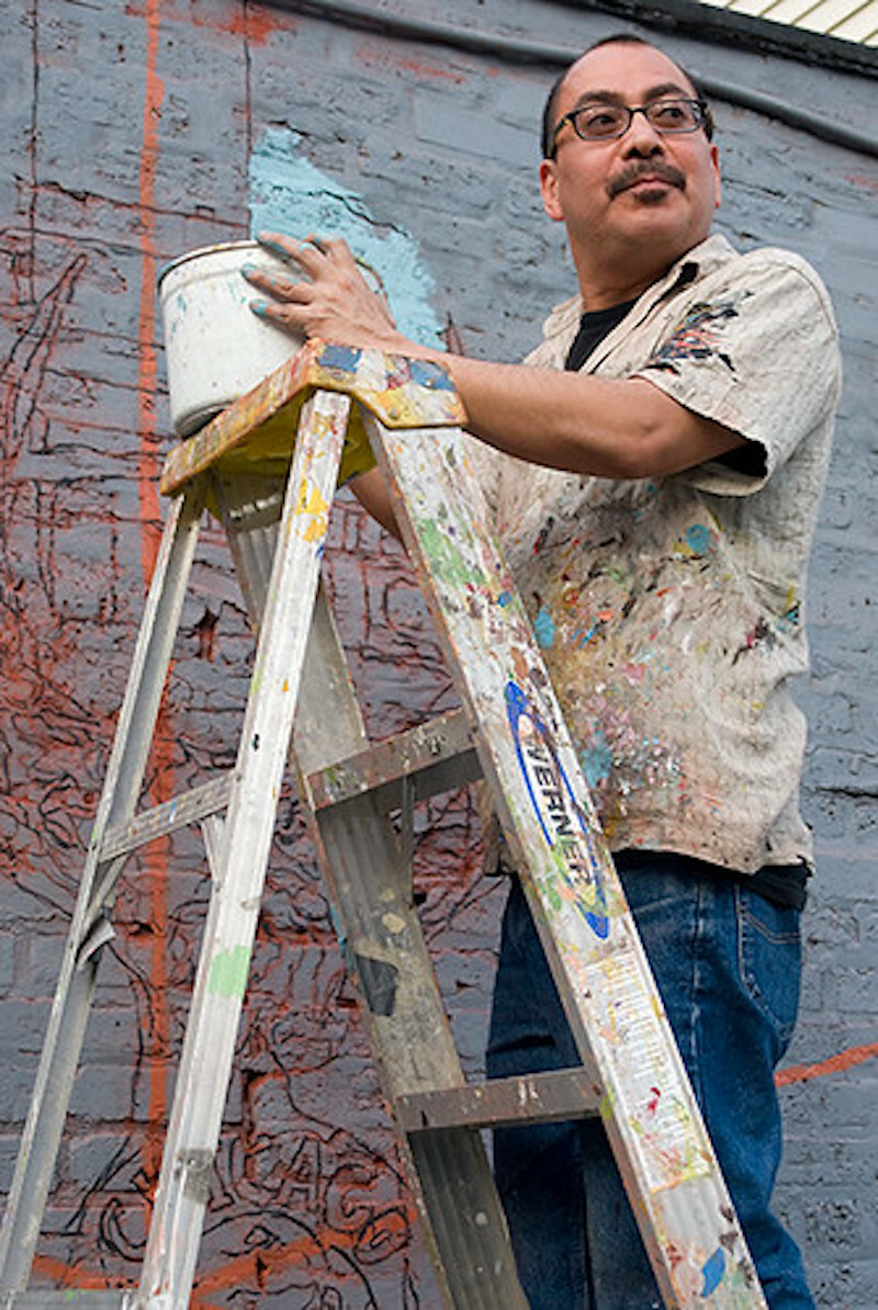 A man stands on a ladder and holds a bucket in front of a brick wall