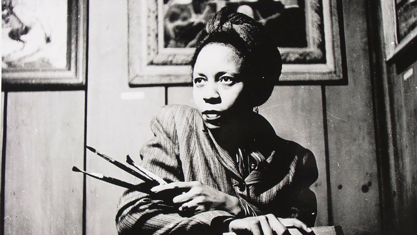 A seated woman crosses her arms and holds paint brushes in her hands