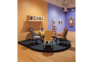 Foreground: An installation of chairs, each with a picture on the chair back, in a circle on a black platform.
