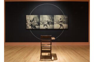 An illuminated classroom desk faces a painted black wall with three still photo frames hanging on it. Hanging photos depict student protestors.