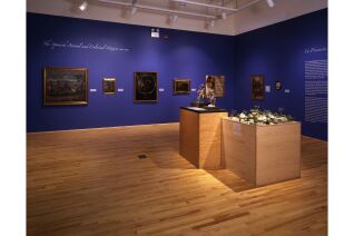 Six casta paintings, a poster, two carved wood figures, and a miniature palenque 3D model pictured from wide angle photograph of gallery