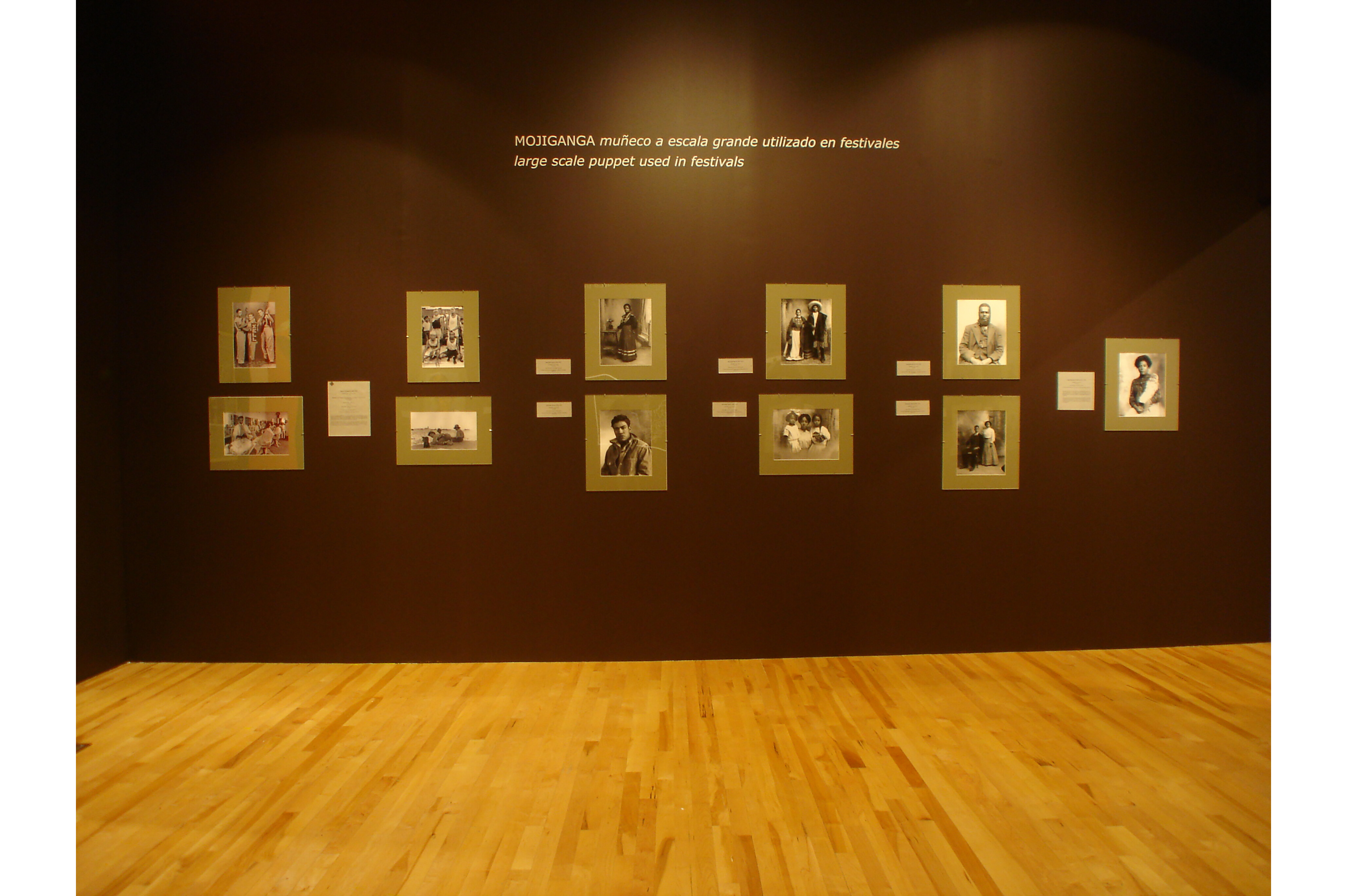 Image of a wall in the gallery with eleven black and white photographs in two rows. Above photographs there is writing. First line reads, “MOHINGA. Muñeco a escala grande utilizado en festivales. Second line reads, “large scale puppet used in festivals”