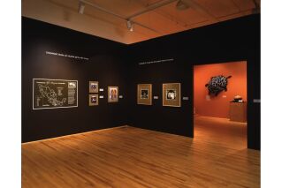 Wide angle photograph of two gallery walls with next room partially visible. Left wall reads, “FANDANGO. Fiesta con musica. Party with music.” Underneath is a poster and three color photographs. Right wall reads, “MORONGA. Longaniza de sangre. Blood sausage.” Below are two black and white photographs. In next room large 3D turtle is visible hanging on wall