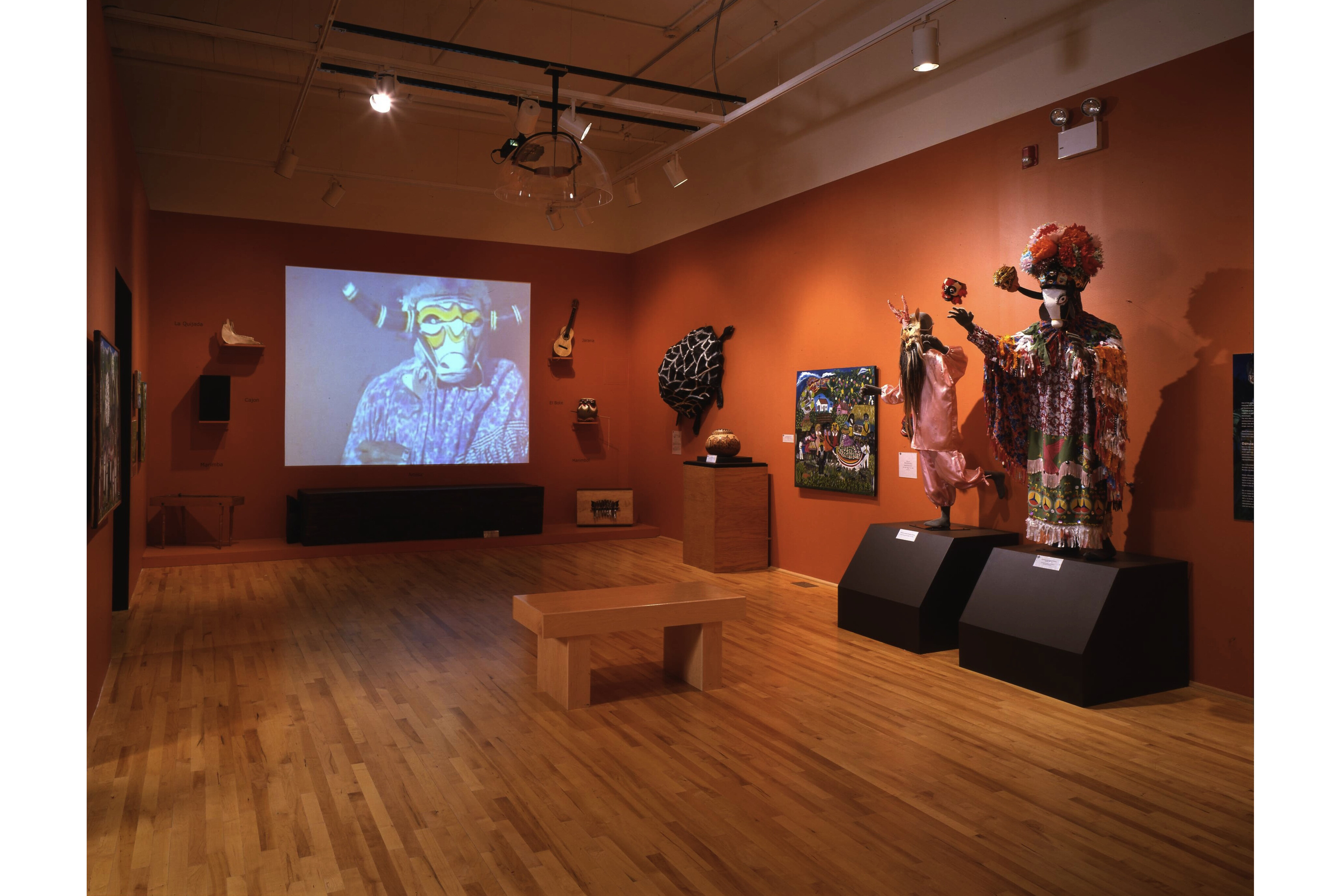 Wide angle photograph of Yanga Carnival artifacts. Instruments, a screen displaying video, a painting, and costumes are visible along the walls