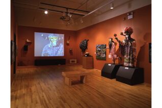 Wide angle photograph of Yanga Carnival artifacts. Instruments, a screen displaying video, a painting, and costumes are visible along the walls