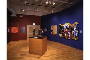 Wide angle photograph shows small figurines in left corner behind glass case, a large painting in right corner, and podium in the center of the room with five sculptures