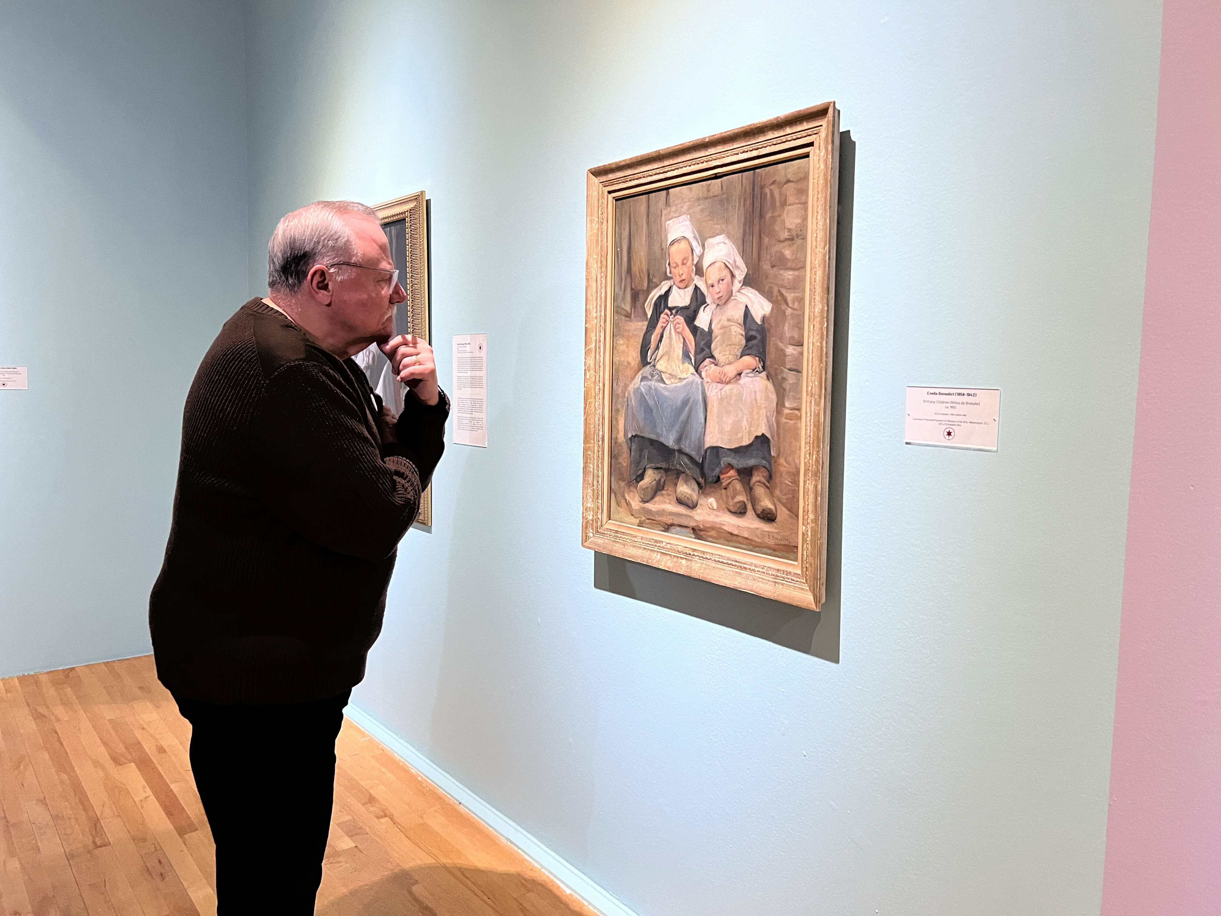 A man leans in as he views a painting of a mother and a daughter. The painting is in a gold frame and hangs on a light blue wall.